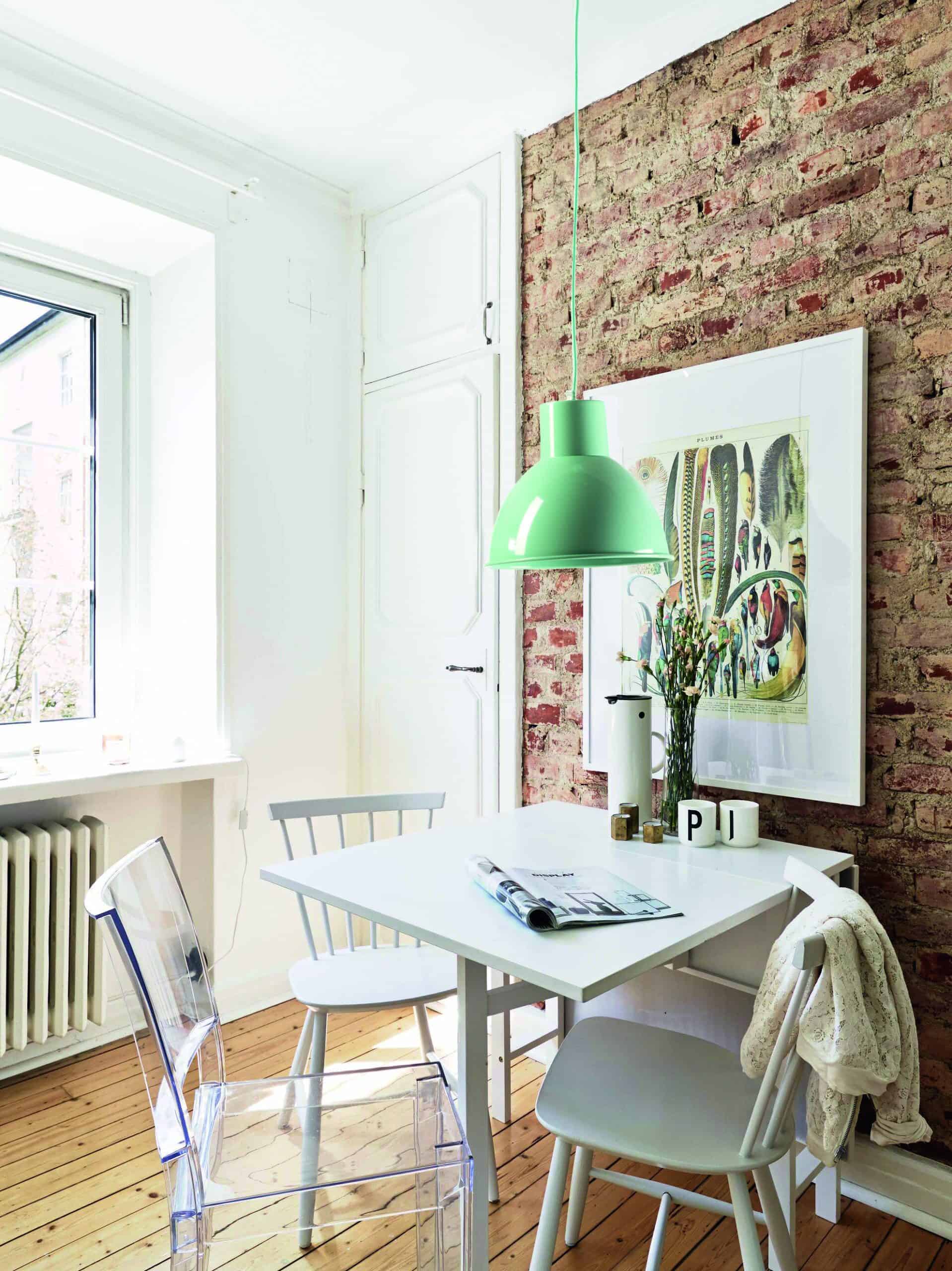 Colorida lámpara de techo verde en comedor con pared de ladrillos y muebles blancos, estilo moderno y natural.